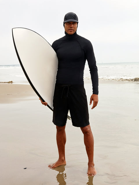 A man wearing a black hooded rashguard carrying a surfboard on the beach