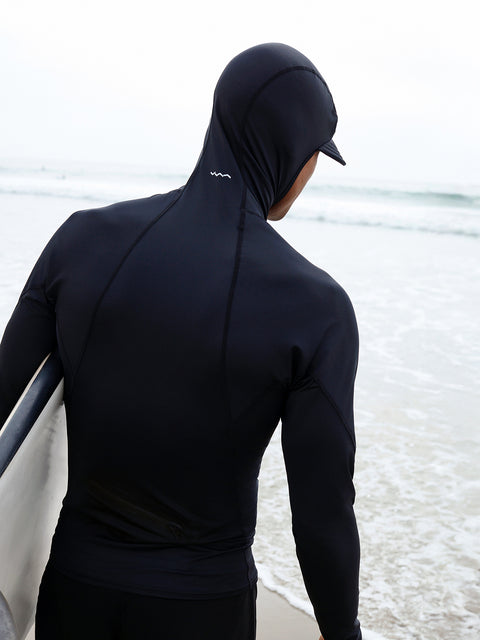 a man wearing a black hooded rash guard carrying a surfboard on the beach