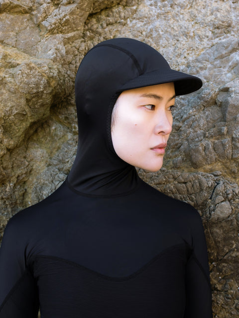 Woman wearing a sun protective black rash guard with a hood for surfing against a stone wall.