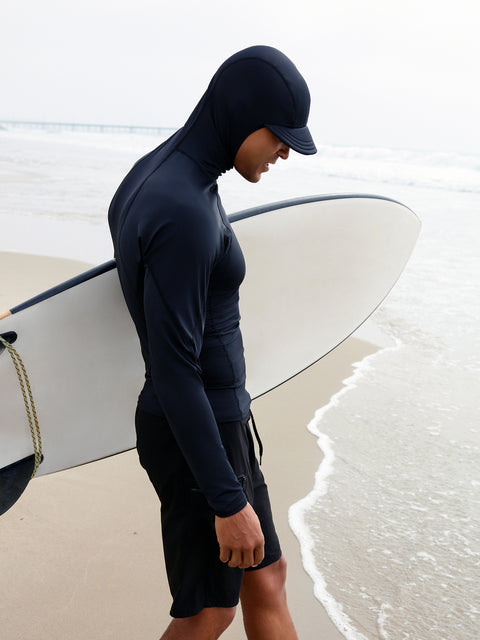 A man wearing a black hooded rashguard with a zippered utility pocket, holding a surfboard on the beach.